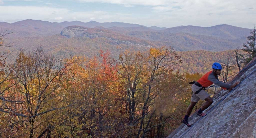 A person wearing safety gear is secured by ropes as they make their way up a rock incline. Behind them is a vast mountainous landscape covered in fall-colored trees. 